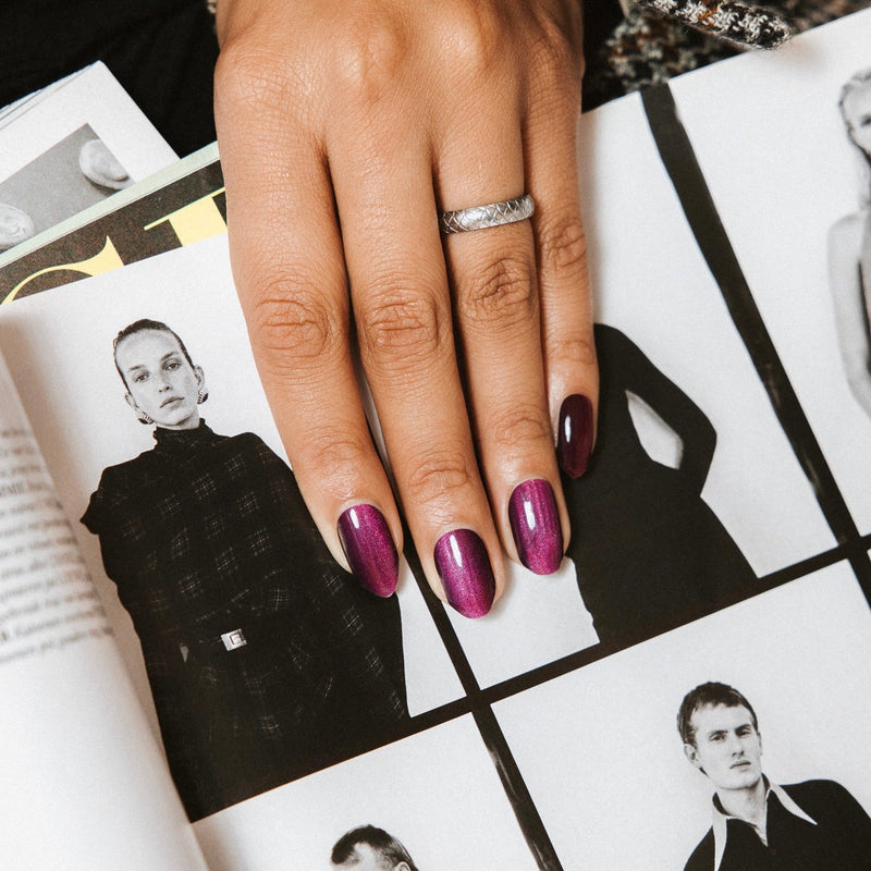 Hand with deep purple-bordeaux shimmer nails resting on fashion magazine pages