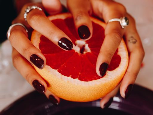 Handen op een halve sinaasappel met donkere rode nagels die de Ruby Red Maniac manicure zijn Ruby Red  maniac Nails Gellak Sticker  Manicure  