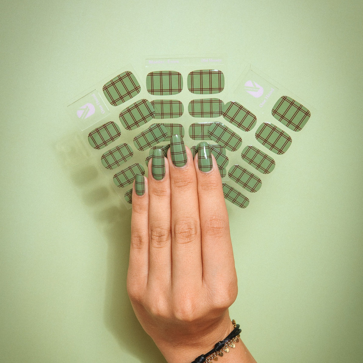 Old Money gel nail sticker sheets by Maniac Nails on a green background, hand with check print nails