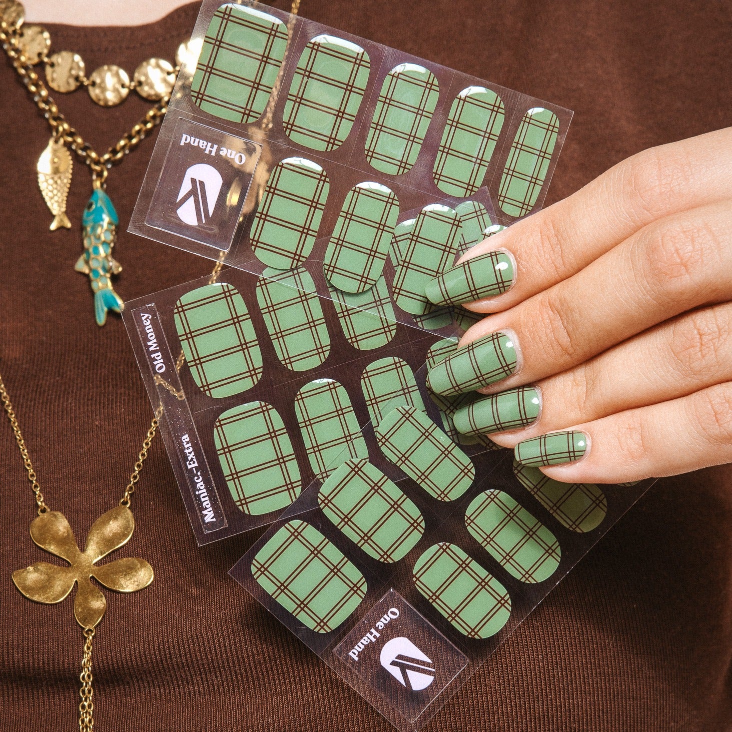 Hand holding Old Money gel nail sticker sheets by Maniac Nails, sage green check nail wraps