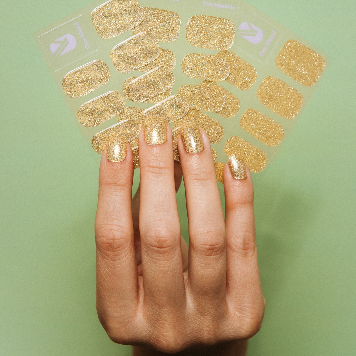 Hand with Unstoppable gold glitter gel nail stickers by Maniac Nails and the sticker sheets