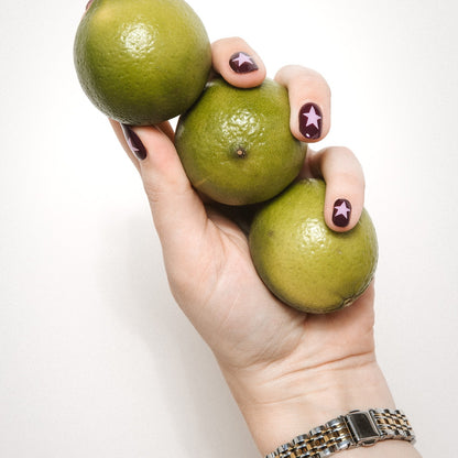 Hand with Wishing Star gel nail stickers by Maniac Nails holding limes, deep plum nails with pink stars